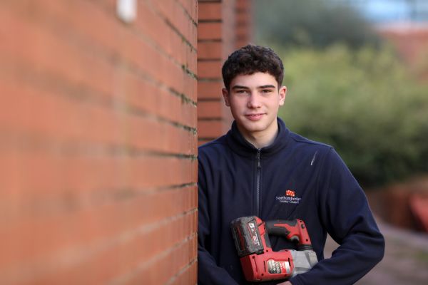 Young man with dark hair standing by a redbrick wall holding a tool