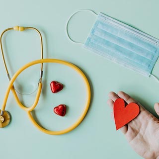 A stethoscope, a face mask are on a table while someone holds a paper heart 