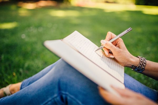 A person sitting on grass writing in a book with a pen