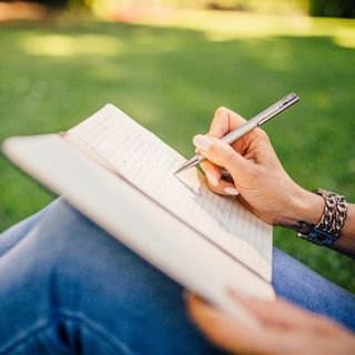 A person sitting on grass writing in a book with a pen
