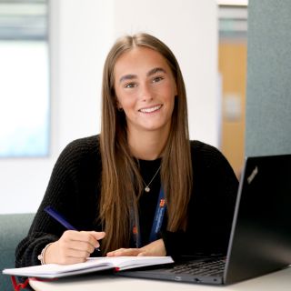 Young woman looking directly at the camera holding a pen, notebook and laptop