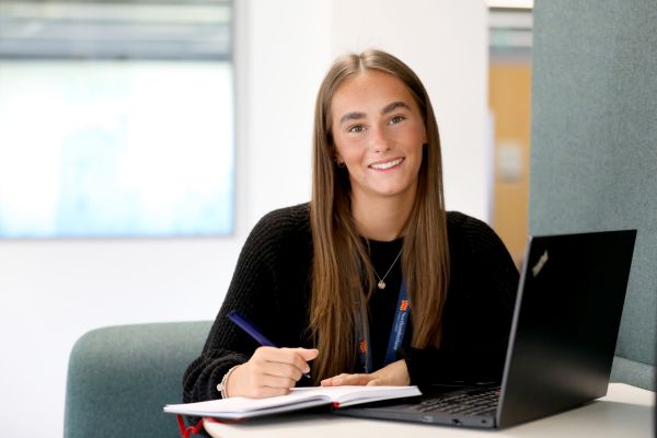 Young woman looking directly at the camera holding a pen, notebook and laptop