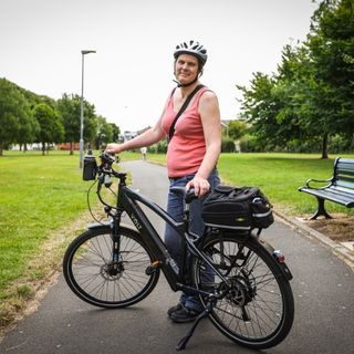 A photograph of Natalie Wright. She wears a white bike helmet and is holding up her e-bike.