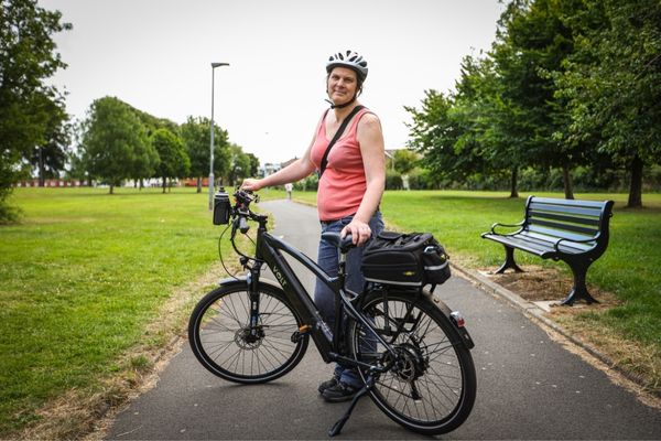 A photograph of Natalie Wright. She wears a white bike helmet and is holding up her e-bike.