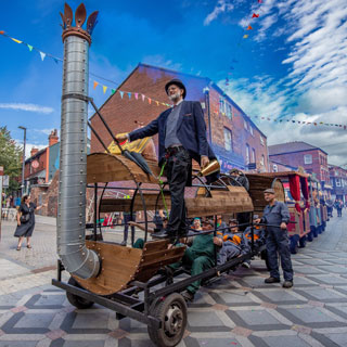 An old train in wheels being drawn through a town centre