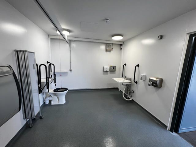 The inside of the accessabile toilet area. A toilet with handles, rise and fall sink unit, adjustable bench, and a hoist.