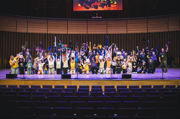 A group of school kids with teachers on a stage holding their music instruments in the air